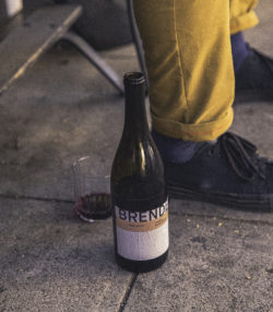 Bottle of Brendel Chorus Cuvee Rouge on sidewalk next to feet and rocks glass