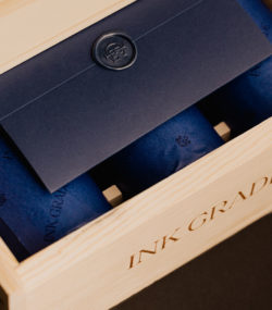 Branded wooden Ink Grade box partially open showing envelope with wax seal and three bottles wrapped in royal blue tissue paper, color