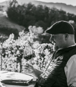 Matt Taylor smiles and swirls a glass of wine at table in vineyard with Ink Grade Howell Mountain Cabernet Sauvignon black and white