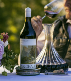 Ink Grade Howell Mountain Cabernet Sauvignon on tablecloth with cork and decanter, winemaker matt taylor sips from a glass in the background
