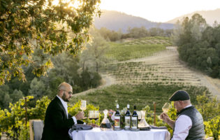 Matt Taylor and Carlton McCoy cheers over table in Ink Grade Vineyard with wine bottles and decanters